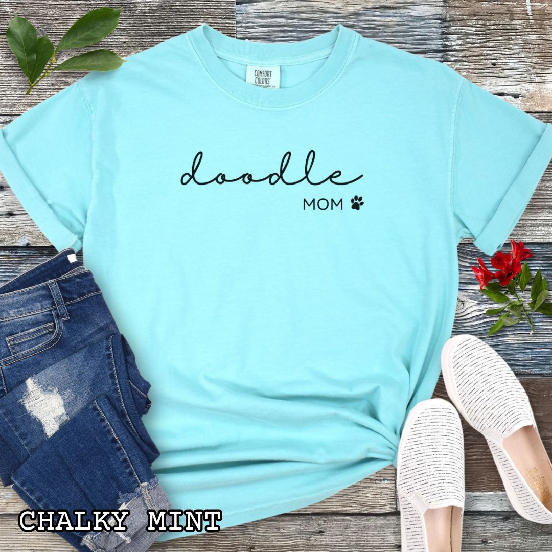 Mint Comfort Colors unisex t-shirt for Doodle dog lovers with 'doodle mom.' text in feminine script and a small paw print, shown on a wooden surface with accessories. Perfect dog mom gift for Goldendoodle, Labradoodle, Aussiedoodle and Bernedoodle moms.