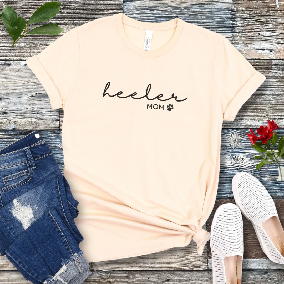 A beige Heeler/Cattle Dog t-shirt with 'heeler mom' written in cursive and a paw print design beneath the text, presented on a wooden surface with a pair of blue jeans and white shoes to the side.