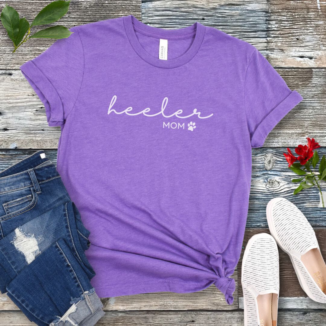 A purple Heeler/Cattle Dog t-shirt with 'heeler mom' written in cursive and a paw print design beneath the text, presented on a wooden surface with a pair of blue jeans and white shoes to the side.