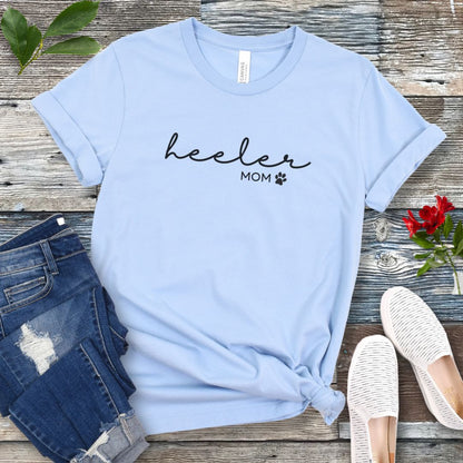 A light blue Heeler/Cattle Dog t-shirt with 'heeler mom' written in cursive and a paw print design beneath the text, presented on a wooden surface with a pair of blue jeans and white shoes to the side.