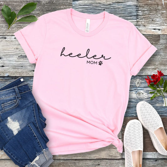 A pink Heeler/Cattle Dog t-shirt with 'heeler mom' written in cursive and a paw print design beneath the text, presented on a wooden surface with a pair of blue jeans and white shoes to the side.