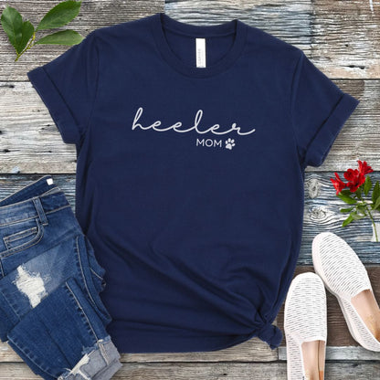 A navy Heeler/Cattle Dog t-shirt with 'heeler mom' written in cursive and a paw print design beneath the text, presented on a wooden surface with a pair of blue jeans and white shoes to the side.