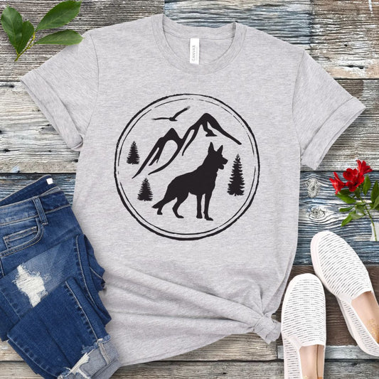 Gray t-shirt with a mountain, trees, and German Shepherd Dog graphic on a wooden surface with jeans and shoes.