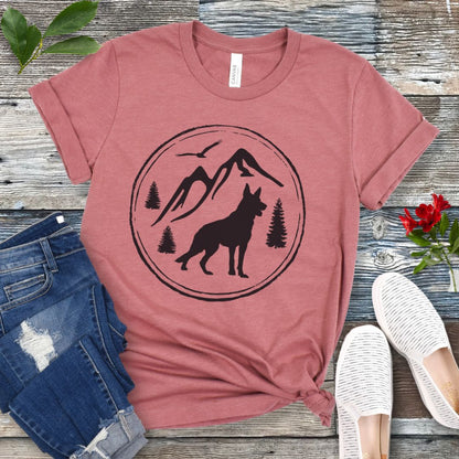 Mauve t-shirt with a mountain, trees, and German Shepherd Dog graphic on a wooden surface with jeans and shoes.