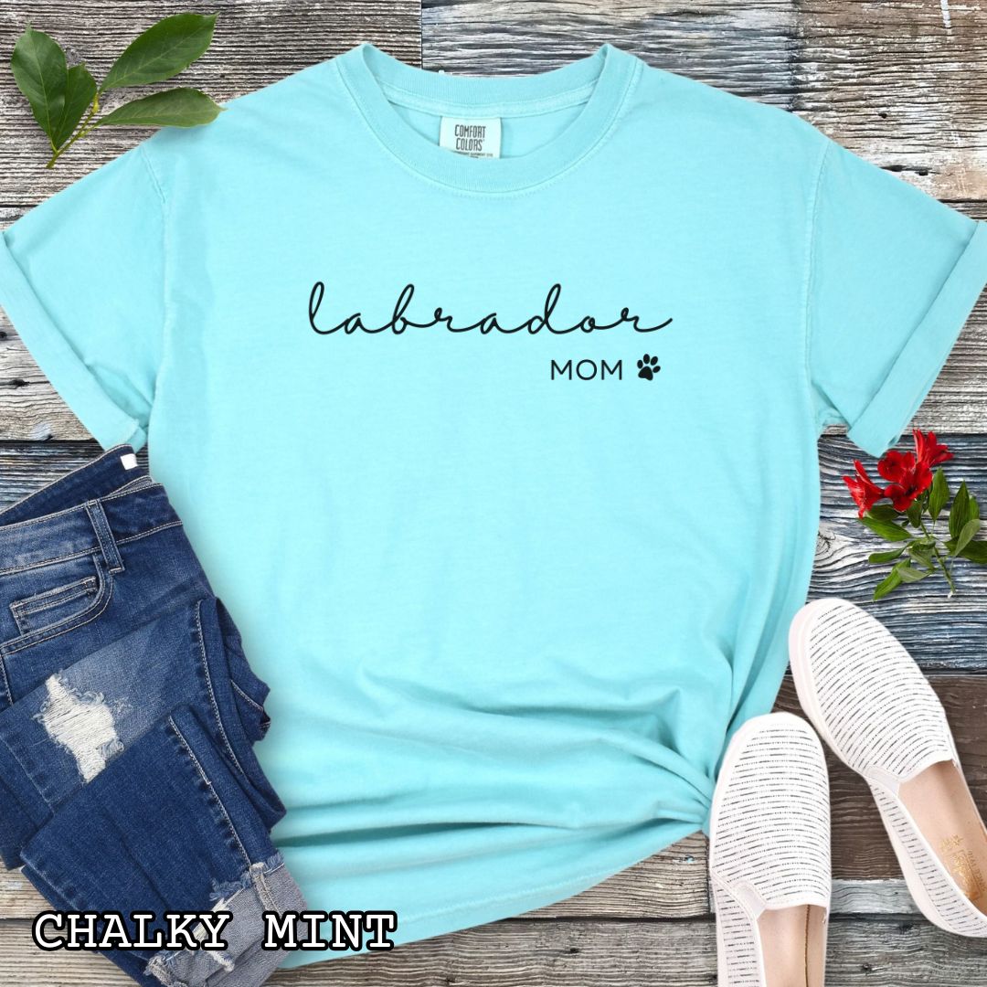 Mint Comfort Colors unisex t-shirt for Labrador dog lovers with 'labrador mom' text in feminine script and a small paw print, shown on a wooden surface with accessories. Perfect dog mom gift for Labrador Retriever moms.
