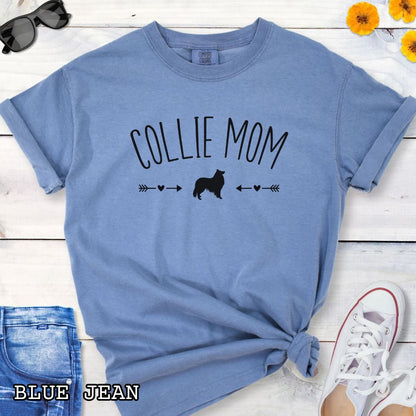 Blue Comfort Colors t-shirt with 'Collie Mom' text in playful lettering and Rough Collie dog graphic accented with small hearts and arrows, shown on a wooden surface with sunglasses and jeans.
