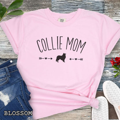 Pink Comfort Colors t-shirt with 'Collie Mom' text in playful lettering and Rough Collie dog graphic accented with small hearts and arrows, shown on a wooden surface with sunglasses and jeans.