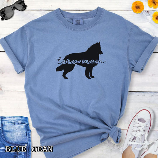 Blue Comfort Colors t-shirt with Belgian Tervuren silhouette and 'Terv Mom' text in handwritten script lettering, shown on a wooden surface with denim jeans and a flower.