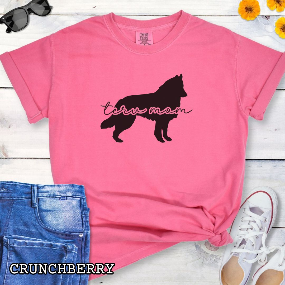 Pink Comfort Colors t-shirt with Belgian Tervuren silhouette and 'Terv Mom' text in handwritten script lettering, shown on a wooden surface with denim jeans and a flower.