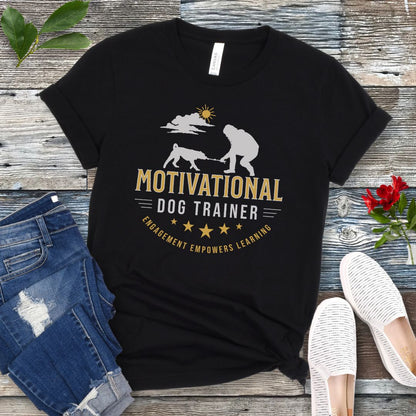 Black t-shirt with 'Motivational Dog Trainer' graphic on a wooden surface with jeans and shoes.