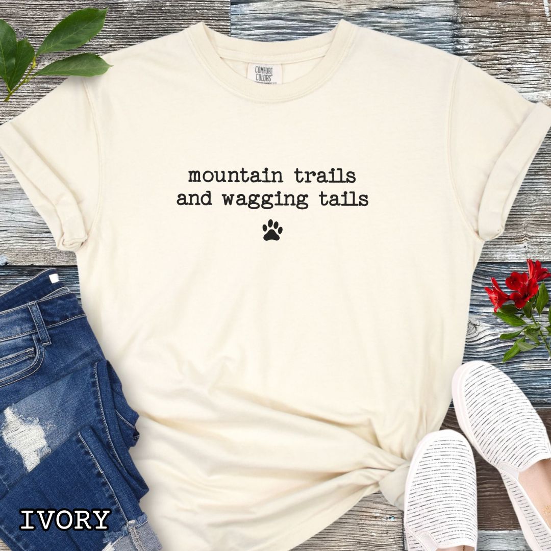 Beige Comfort Colors t-shirt for outdoorsy dog lovers with 'mountain trails and wagging tails' text in typewriter font and a small paw print accent, shown on a wooden surface with shoes and jeans.