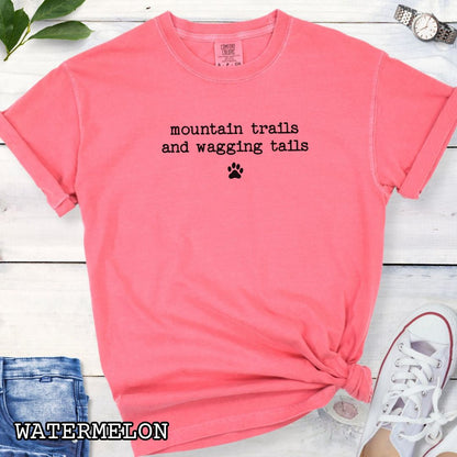 Pink Comfort Colors t-shirt for outdoorsy dog lovers with 'mountain trails and wagging tails' text in typewriter font and a small paw print accent, shown on a wooden surface with shoes and jeans.