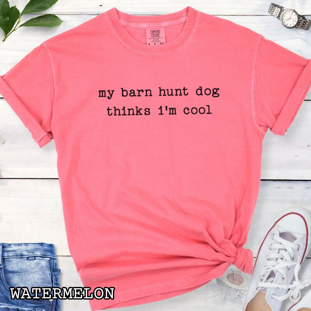 Pink Comfort Colors dog sports t-shirt with text 'my barn hunt dog thinks i'm cool' on a wooden surface with sunglasses and jeans.
