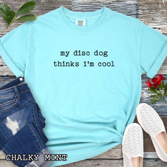 Mint Comfort Colors dog sports t-shirt with text 'my disc dog thinks i'm cool' on a wooden surface with jeans and shoes.
