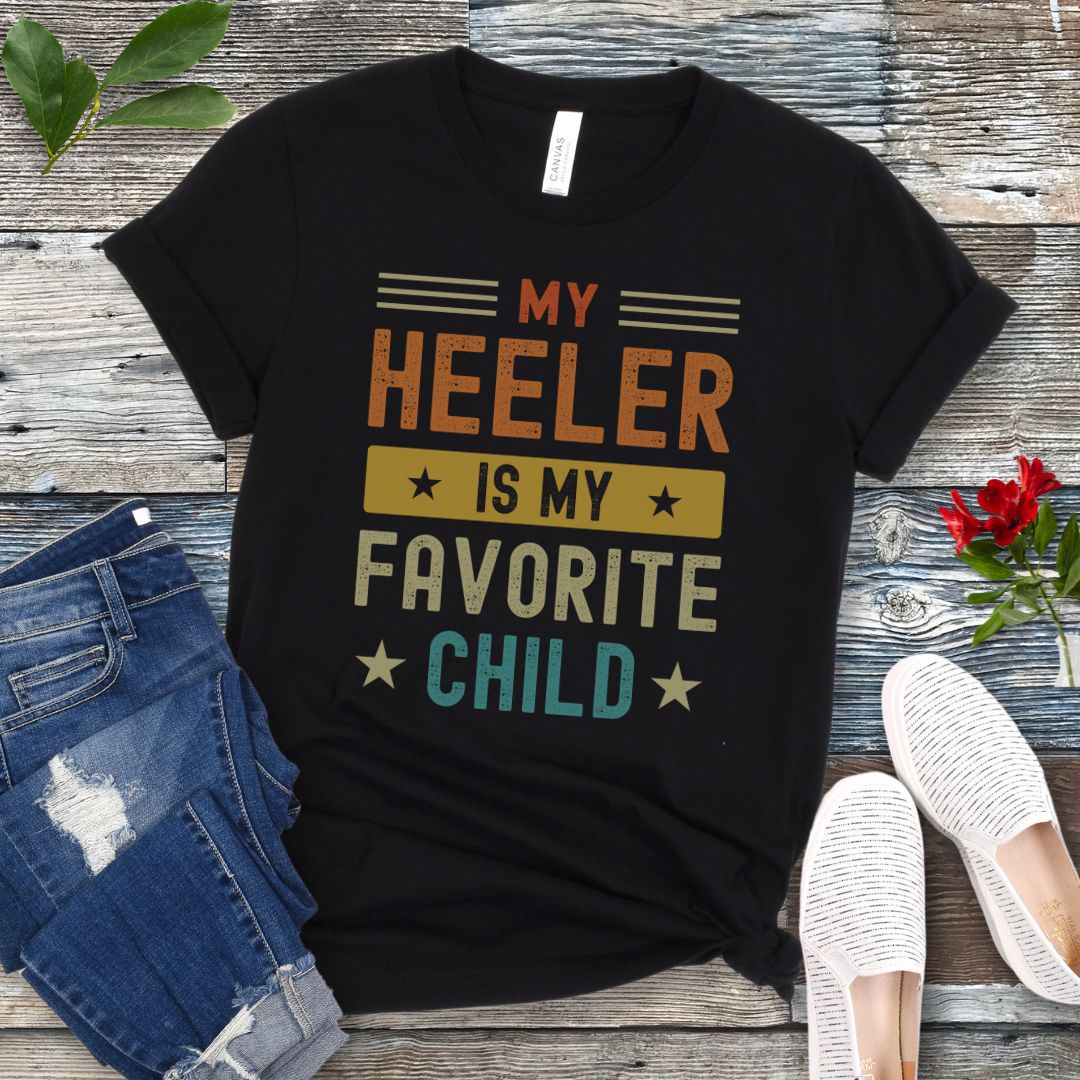 Black dog lover t-shirt with the text 'MY HEELER IS MY FAVORITE CHILD' printed in orange and yellow, placed on a wooden surface with a pair of blue jeans and white shoes in the background.