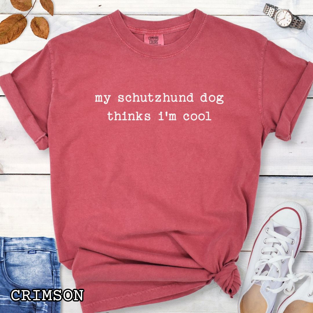 Red Comfort Colors IGP t-shirt with text 'my schutzhund dog thinks i'm cool' on a wooden surface with jeans and sneakers.