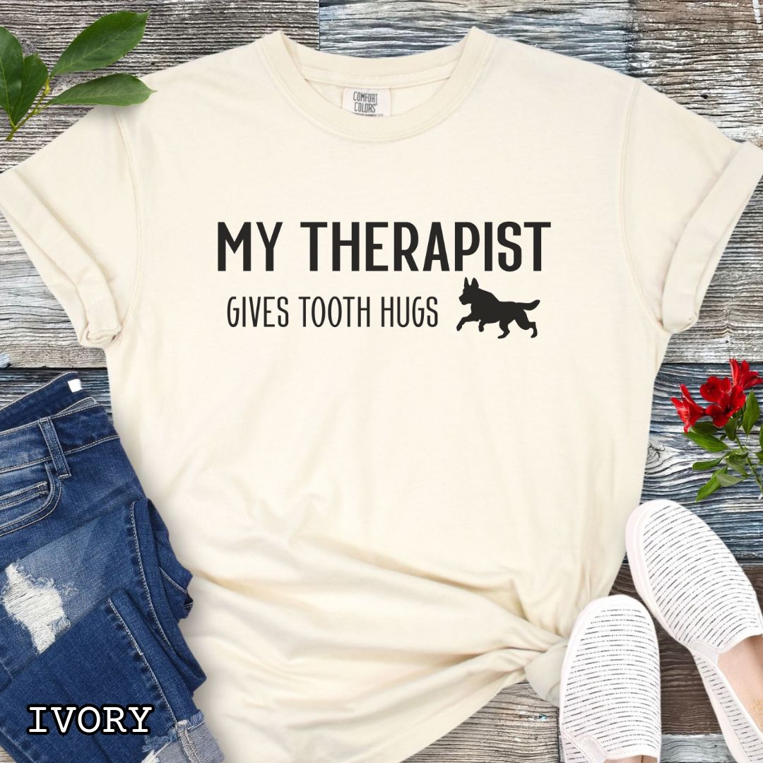 Ivory Comfort Colors t-shirt for bitework and protection dog trainers with humorous text 'My Therapist Gives Tooth Hugs' and dog graphic on a wooden surface.