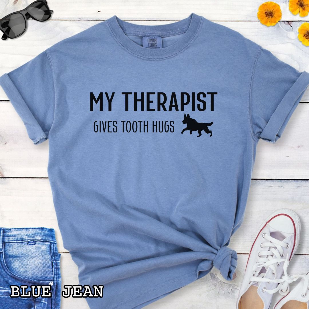 Blue Comfort Colors t-shirt for bitework and protection dog trainers with humorous text 'My Therapist Gives Tooth Hugs' and dog graphic on a wooden surface.