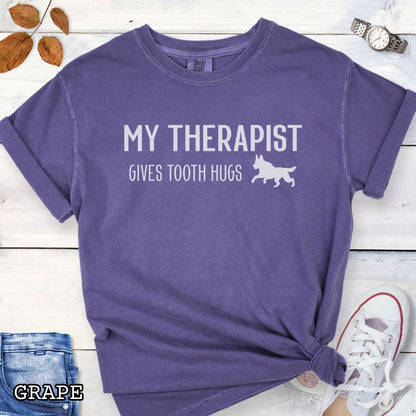 Purple Comfort Colors t-shirt for bitework and protection dog trainers with humorous text 'My Therapist Gives Tooth Hugs' and dog graphic on a wooden surface.