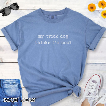 Blue Comfort Colors t-shirt with 'my trick dog thinks i'm cool' text on a wooden surface with sunglasses and jeans.