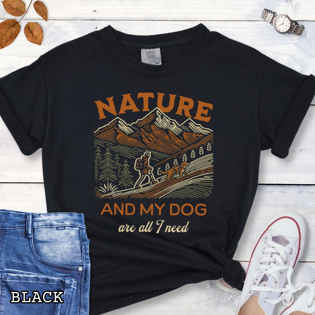 Black Comfort Colors t-shirt with graphic of mountains, hiker and dog and 'Nature and My Dog are all I need' text, shown on a wooden surface with jeans and sneakers.