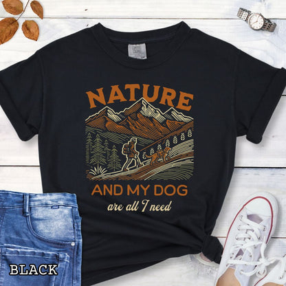 Black Comfort Colors t-shirt with graphic of mountains, hiker and dog and 'Nature and My Dog are all I need' text, shown on a wooden surface with jeans and sneakers.