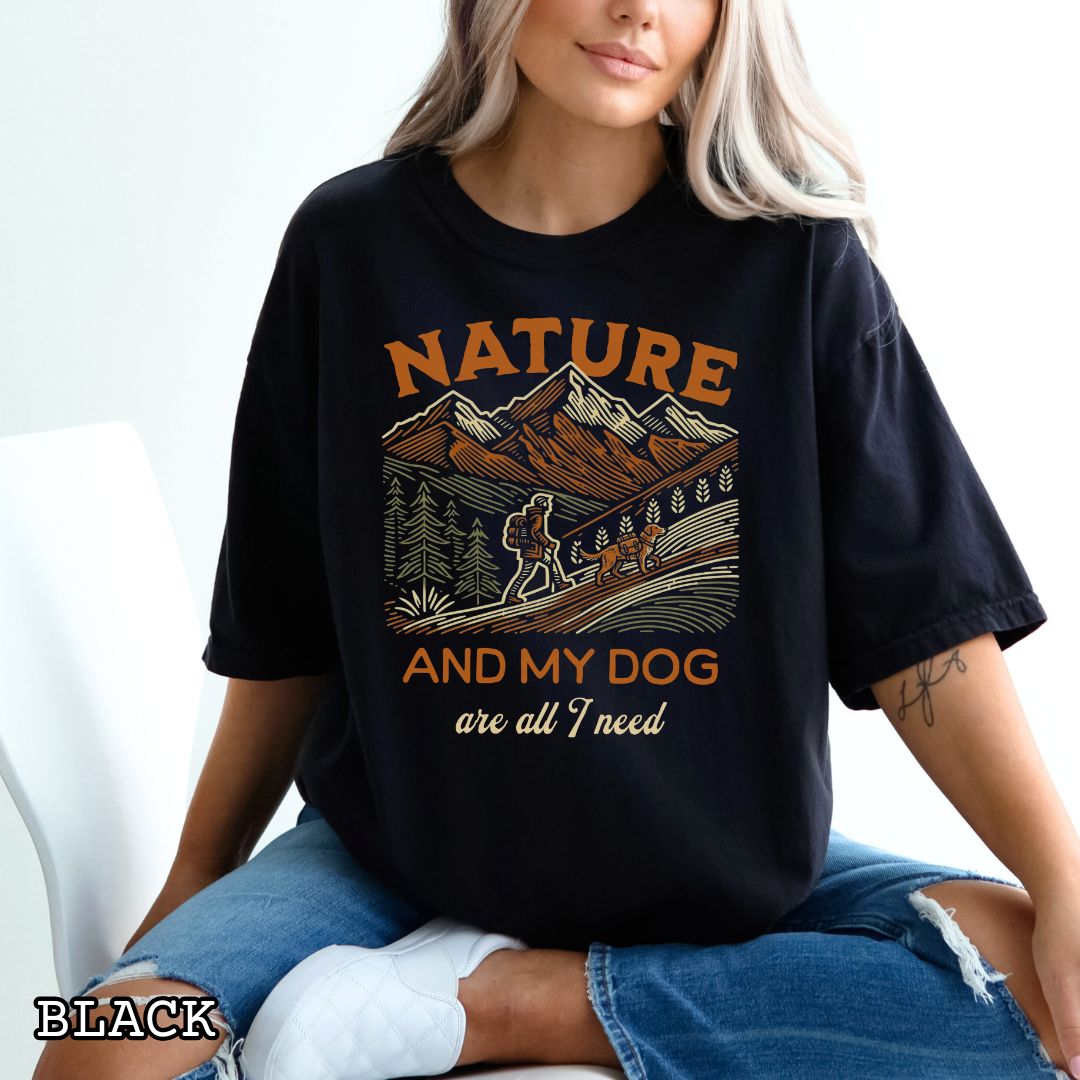 Person wearing a black Comfort Colors t-shirt with graphic of mountains, hiker and dog and 'Nature and My Dog are all I need' text.