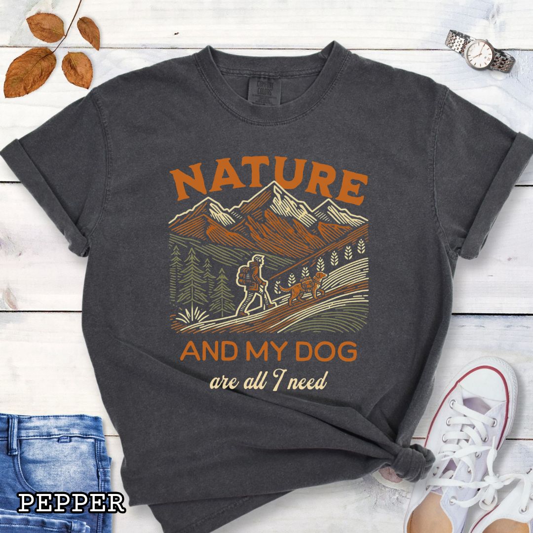 Dark grey Comfort Colors t-shirt with graphic of mountains, hiker and dog and 'Nature and My Dog are all I need' text, shown on a wooden surface with jeans and sneakers.