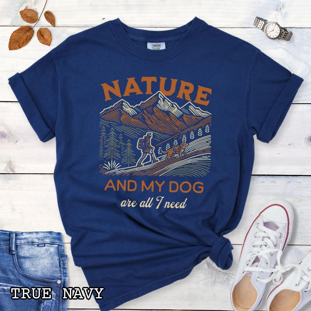 Navy blue Comfort Colors t-shirt with graphic of mountains, hiker and dog and 'Nature and My Dog are all I need' text, shown on a wooden surface with jeans and sneakers.
