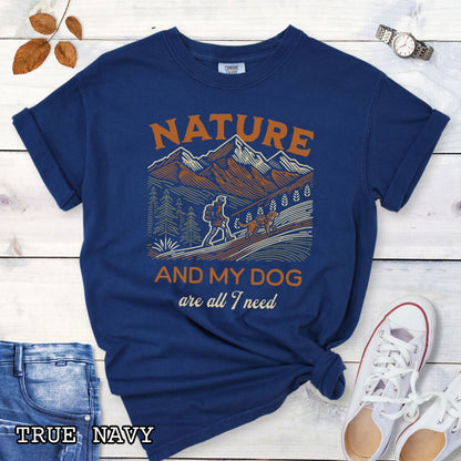 Navy blue Comfort Colors t-shirt with graphic of mountains, hiker and dog and 'Nature and My Dog are all I need' text, shown on a wooden surface with jeans and sneakers.