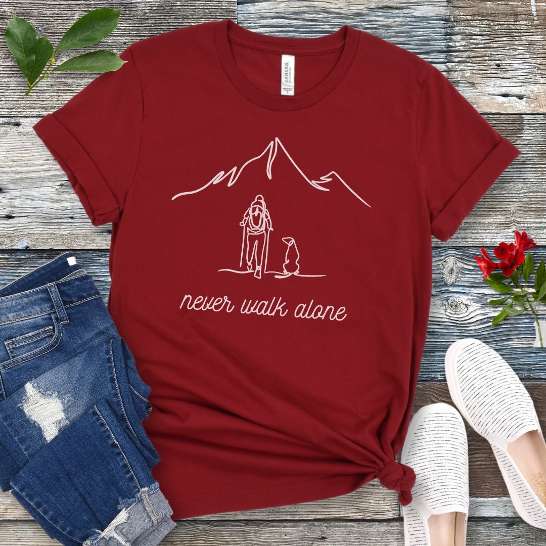 A dark red dog lover hiking t-shirt with a line art drawing of a person with a dog walking toward a mountain, and the text 'never walk alone', displayed on a wooden surface with a pair of jeans and white shoes in the background.