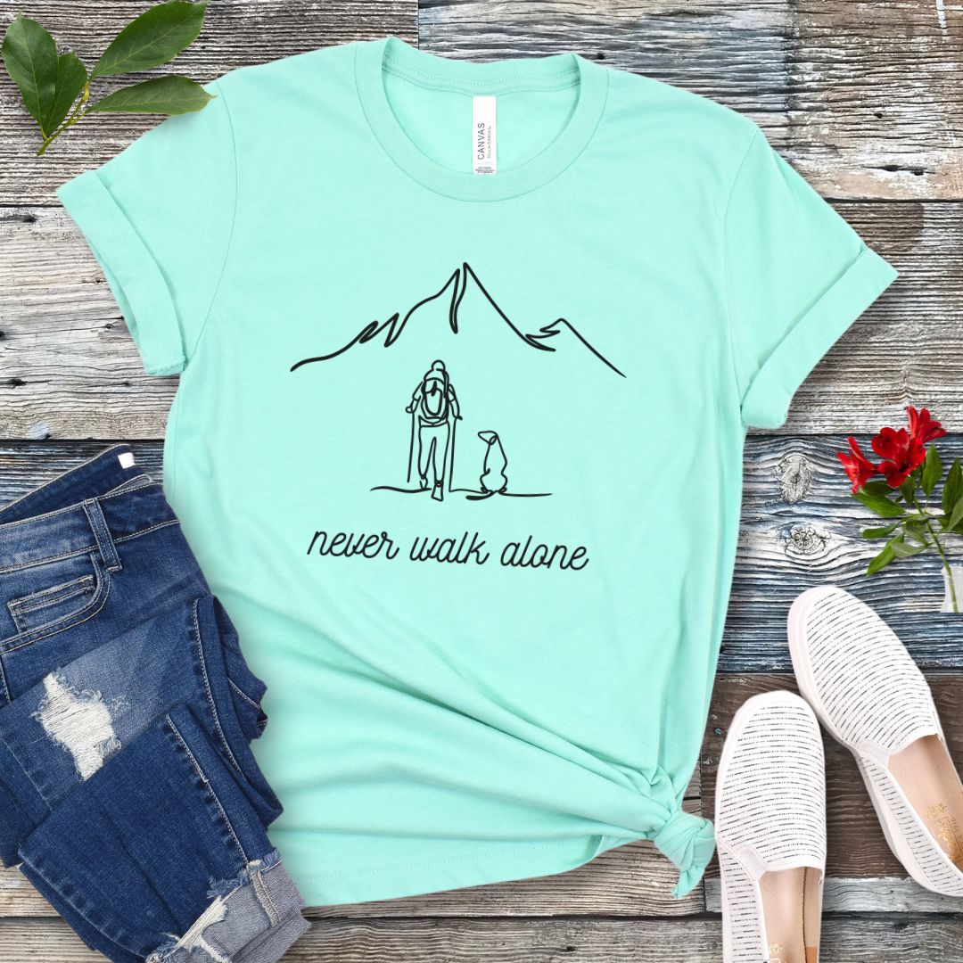 A mint green dog lover hiking t-shirt with a line art drawing of a person with a dog walking toward a mountain, and the text 'never walk alone', displayed on a wooden surface with a pair of jeans and white shoes in the background.