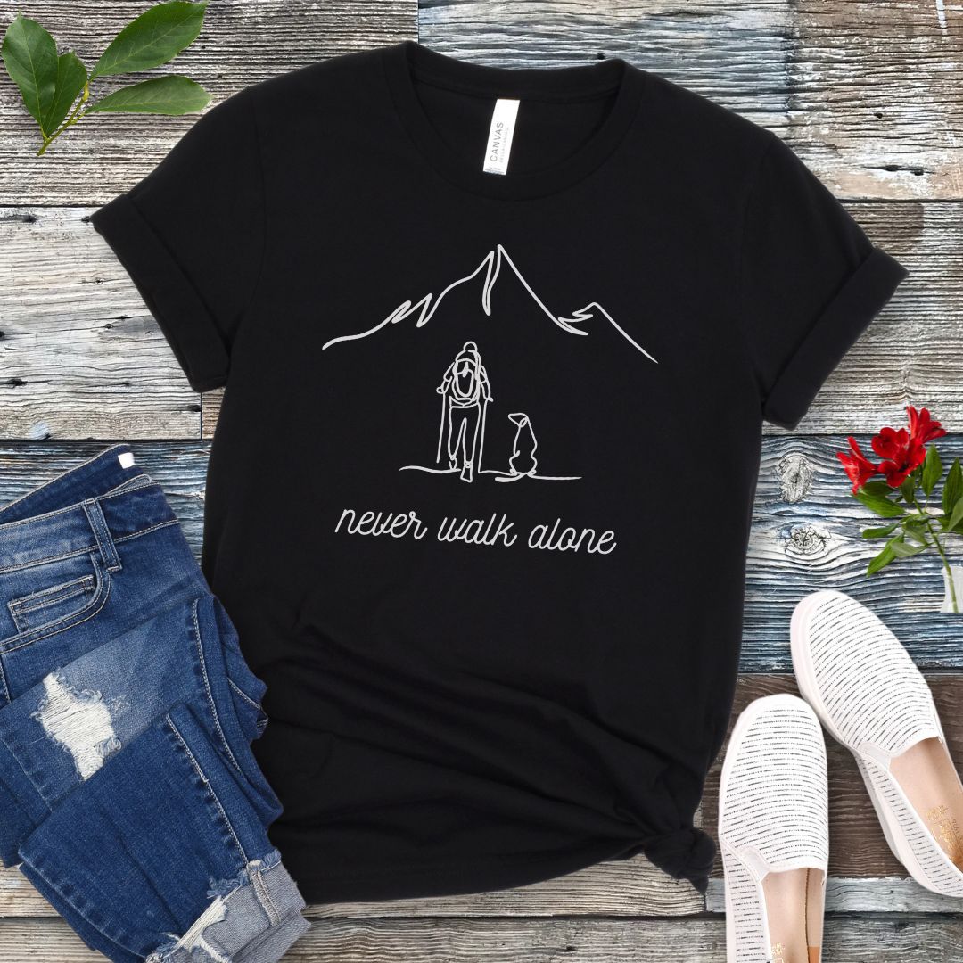 A black dog lover hiking t-shirt with a line art drawing of a person with a dog walking toward a mountain, and the text 'never walk alone', displayed on a wooden surface with a pair of jeans and white shoes in the background.