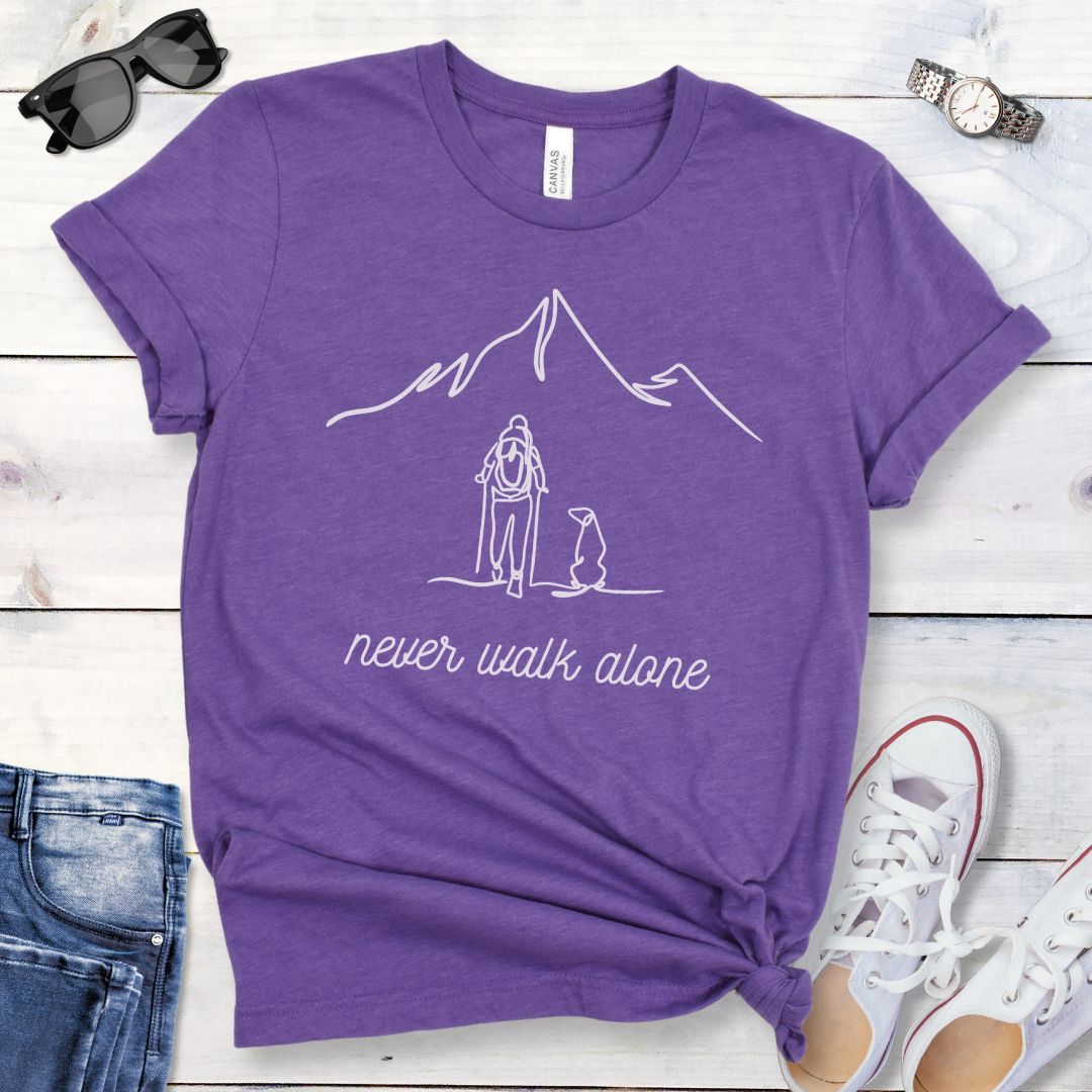 A purple dog lover hiking t-shirt with a line art drawing of a person with a dog walking toward a mountain, and the text 'never walk alone', displayed on a wooden surface with a pair of jeans and white shoes in the background.
