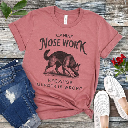 Mauve t-shirt with black text and vintage style dog illustration, stating 'CANINE NOSE WORK BECAUSE MURDER IS WRONG', displayed on a wooden background with denim jeans and white shoes.