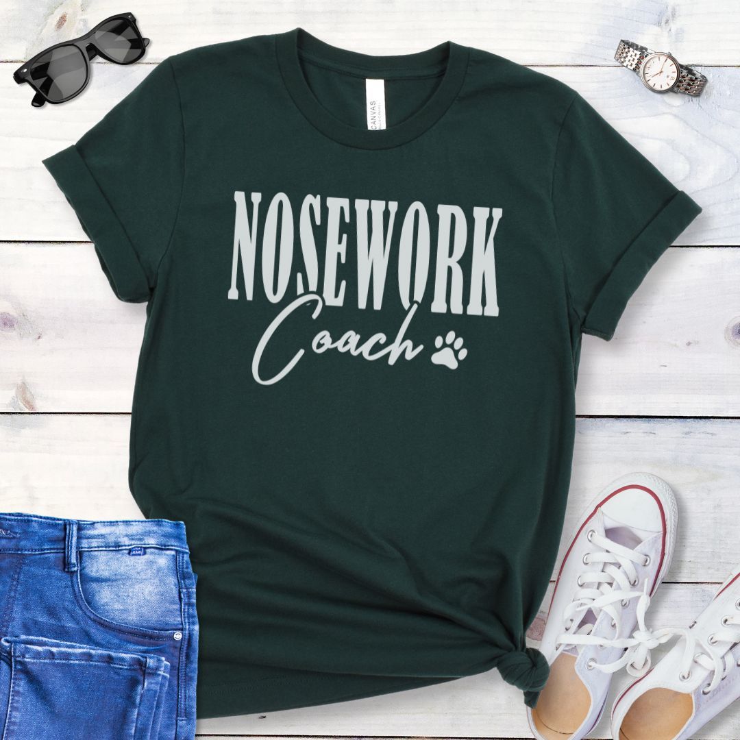 Forest green dog trainer t-shirt with 'Nosework Coach' text, presented on a wooden surface with jeans and shoes.