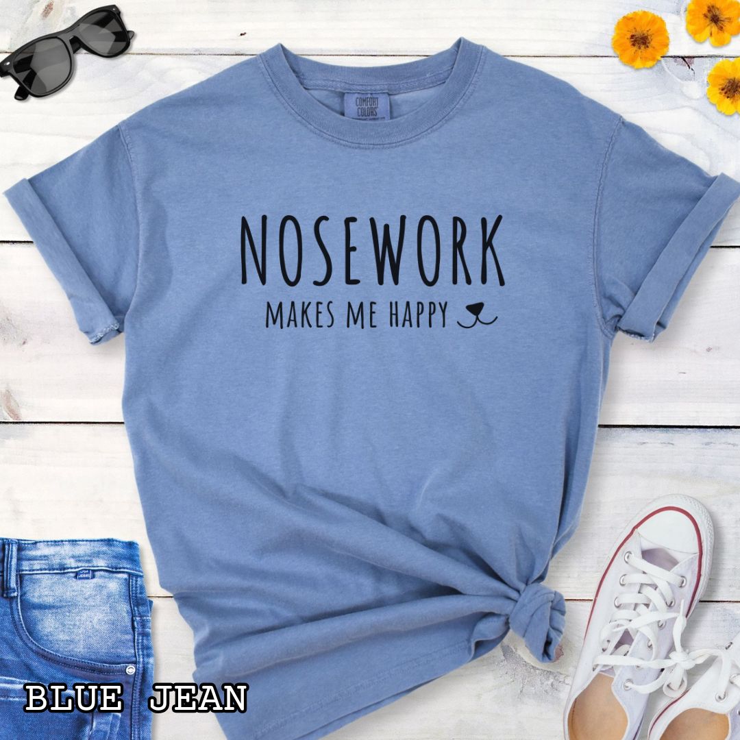 Blue Comfort Colors t-shirt for dog scent trainers with 'NOSEWORK makes me happy' text and a dog nose accent, shown on a wooden surface with sunglasses, jeans, and sneakers.