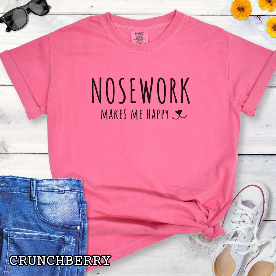 Pink Comfort Colors t-shirt for dog scent trainers with 'NOSEWORK makes me happy' text and a dog nose accent, shown on a wooden surface with sunglasses, jeans, and sneakers.