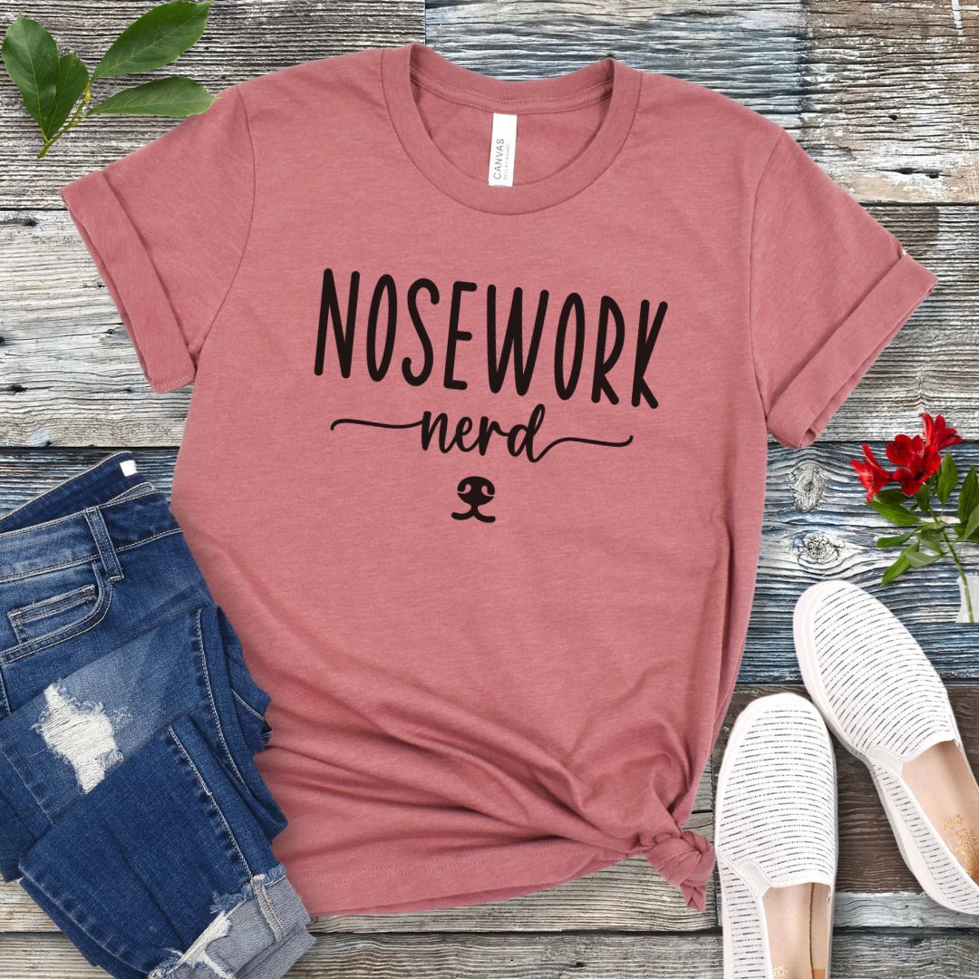 Mauve dog sports t-shirt with 'Nosework nerd' text on a wooden surface with jeans and shoes.