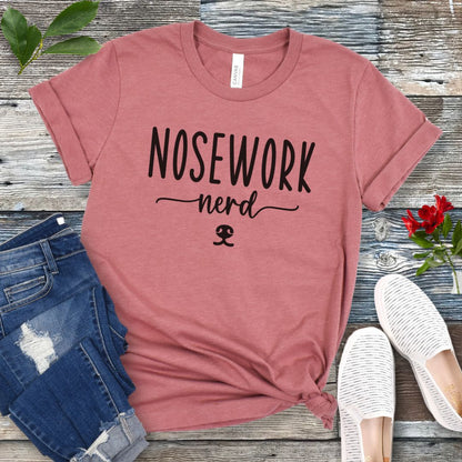 Mauve dog sports t-shirt with 'Nosework nerd' text on a wooden surface with jeans and shoes.