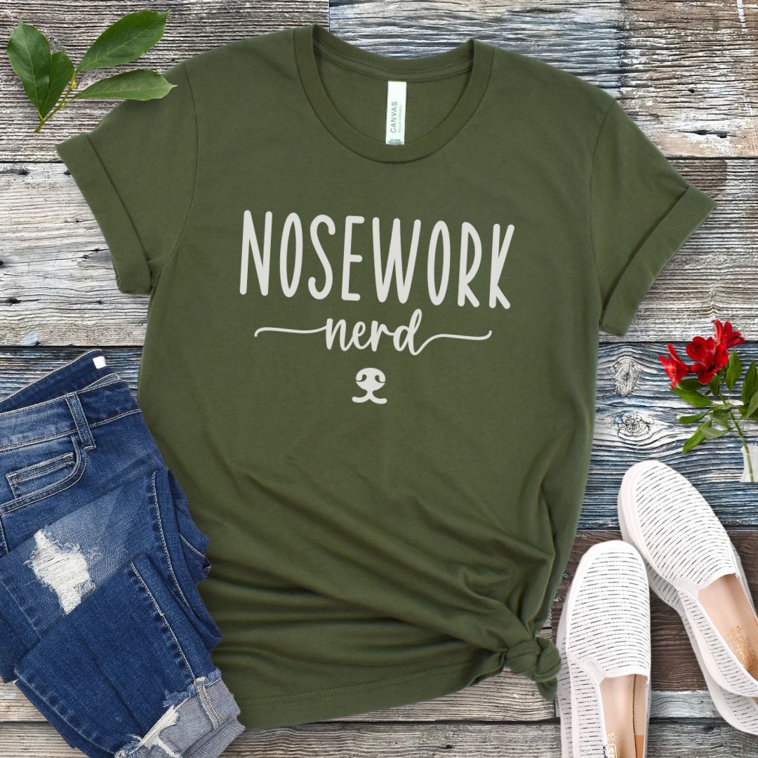Military green dog sports t-shirt with 'Nosework nerd' text on a wooden surface with jeans and shoes.