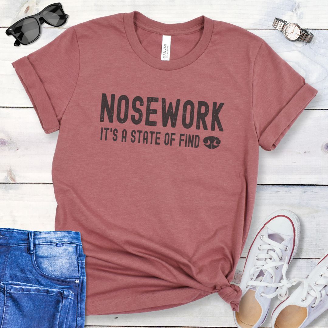 Mauve dog scent training t-shirt with humorous text 'NOSEWORK, it's a state of find' and the graphic of a dog's nose, shown on a wooden surface with jeans and shoes. 