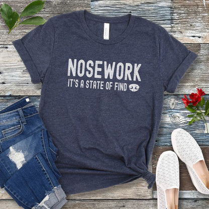 Heather navy dog sports t-shirt with 'NOSEWORK, it's a state of find' text on a wooden surface with jeans and shoes. 