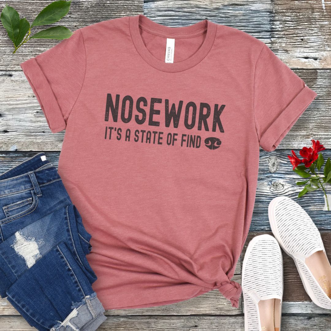 Mauve dog sports t-shirt with 'NOSEWORK, it's a state of find' text on a wooden surface with jeans and shoes. 
