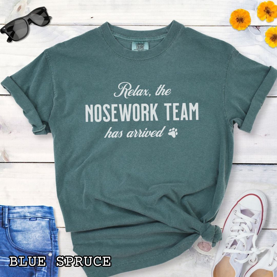 Green Comfort Colors dog sports t-shirt with text 'Relax, the NOSEWORK TEAM has arrived' on a wooden surface with denim jeans and white shoes.