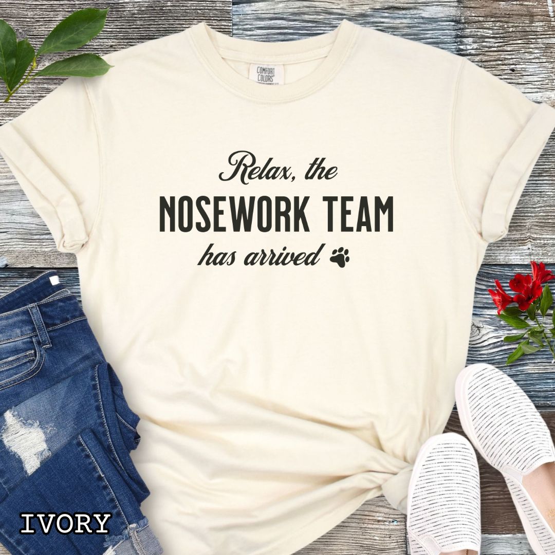 Beige Comfort Colors dog sports t-shirt with text 'Relax, the NOSEWORK TEAM has arrived' on a wooden surface with denim jeans and white shoes.