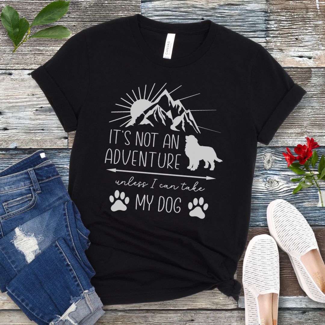 A black dog lover T-shirt with a graphic that says 'IT'S NOT AN ADVENTURE UNLESS I CAN TAKE MY DOG' accompanied by images of a mountain, a dog and paw prints. The shirt is displayed alongside a pair of jeans and a pair of white shoes on a wooden background.