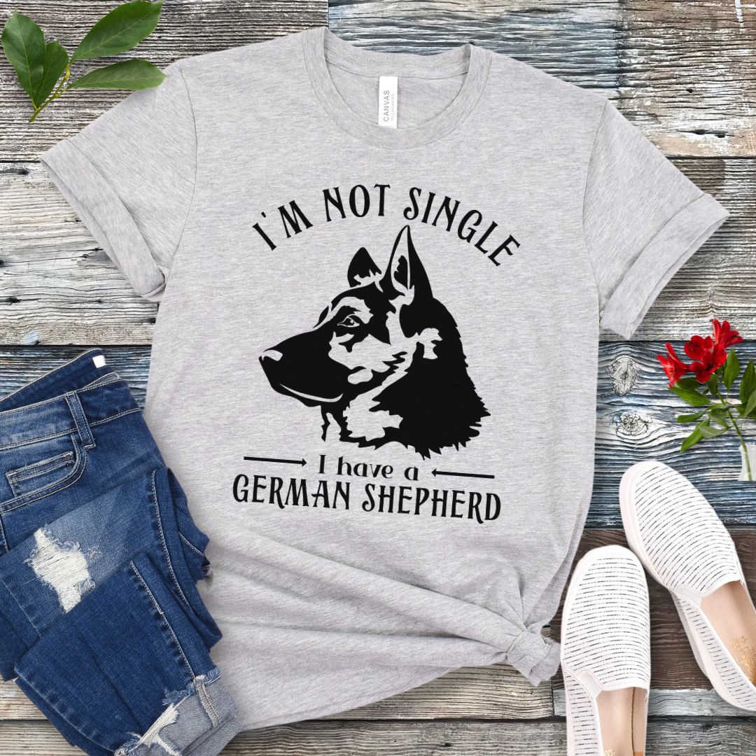 Light grey t-shirt with a German Shepherd graphic and 'I'm Not Single, I have a German Shepherd' text, presented on a wooden surface with jeans and shoes.