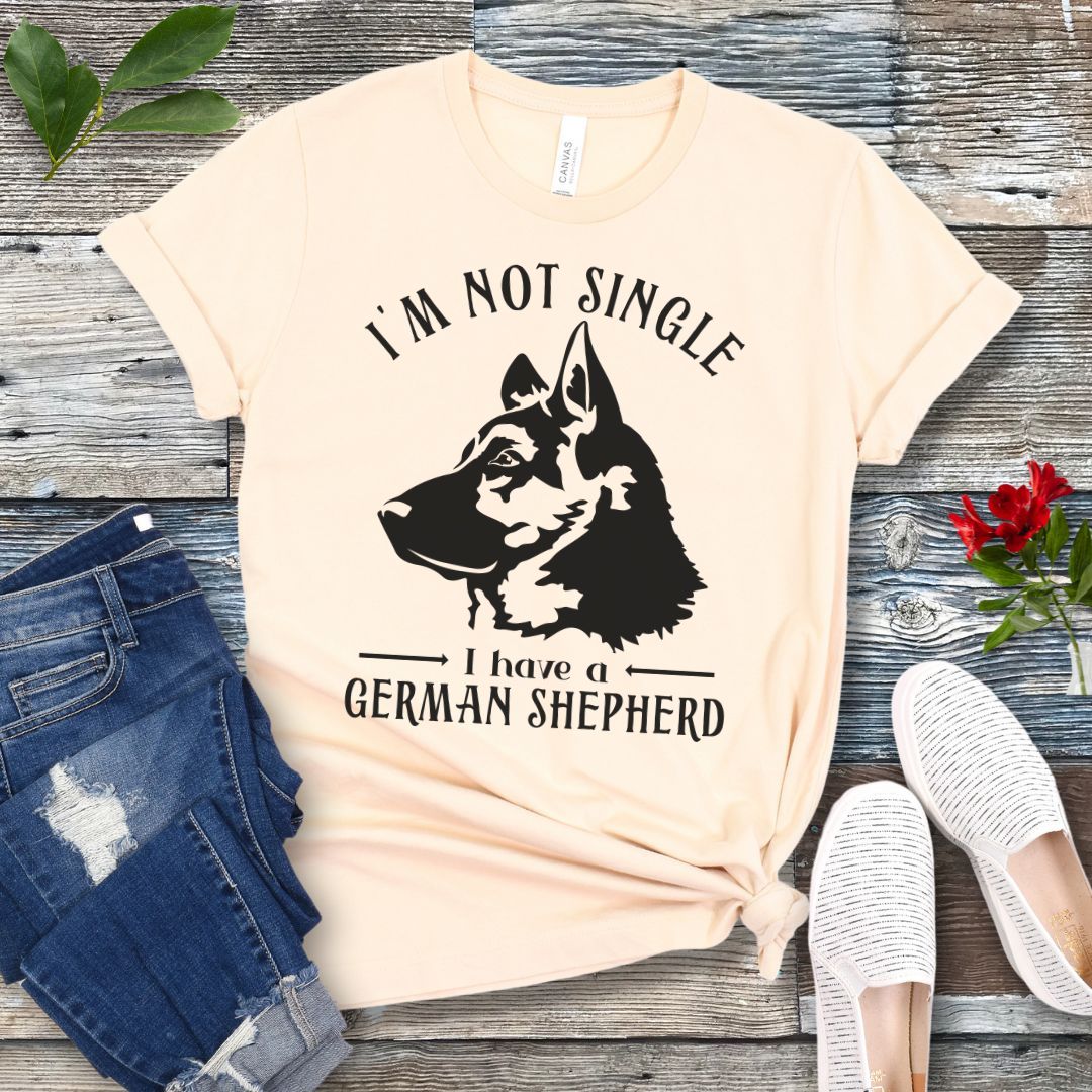 Beige t-shirt with a German Shepherd graphic and 'I'm Not Single, I have a German Shepherd' text, presented on a wooden surface with jeans and shoes.