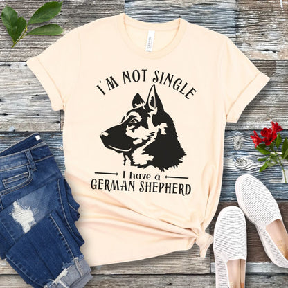 Beige t-shirt with a German Shepherd graphic and 'I'm Not Single, I have a German Shepherd' text, presented on a wooden surface with jeans and shoes.